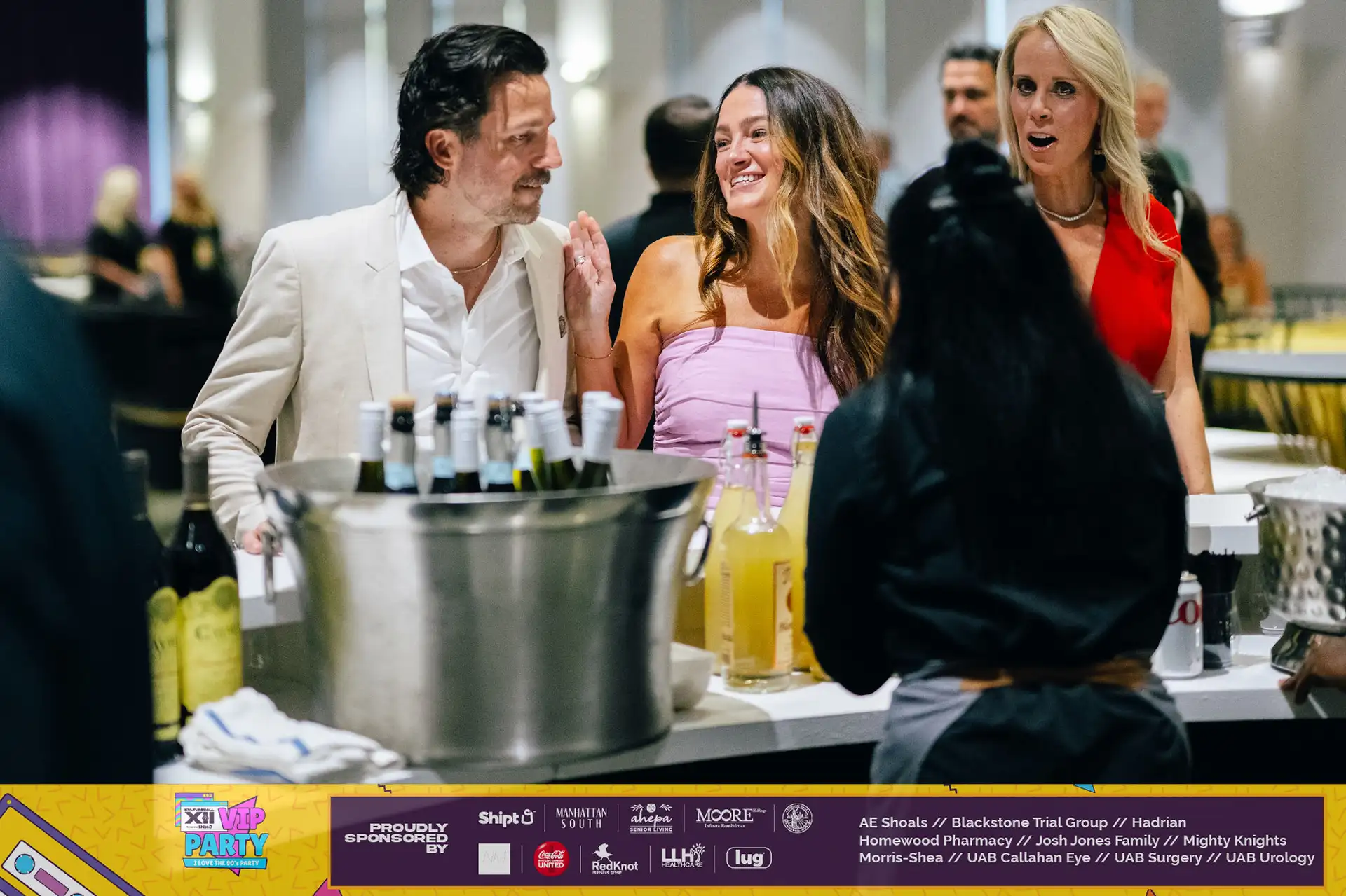 A group of people standing around a table with a variety of drinks, including lemonade, and wine glasses.