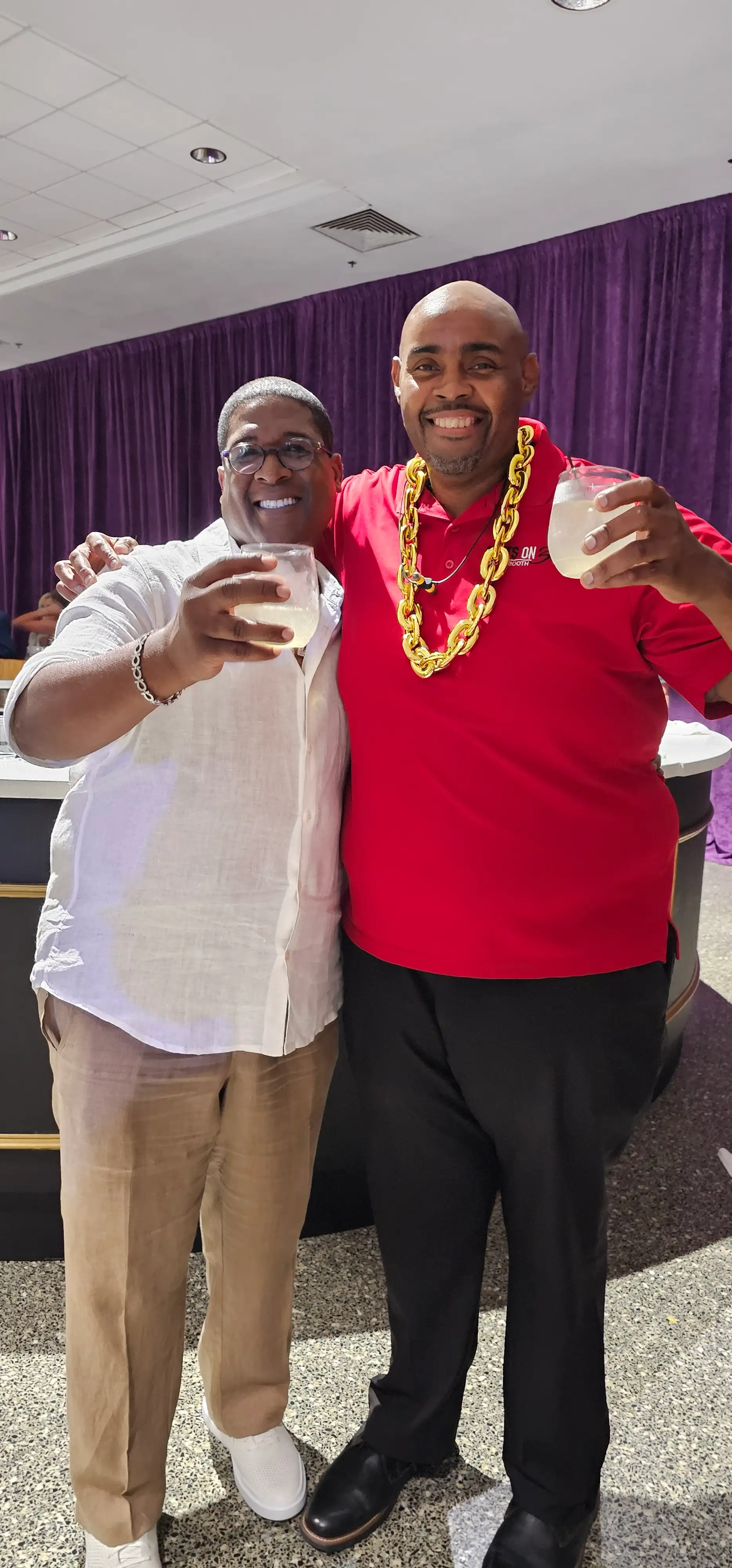 Two men standing next to each other, one holding a glass of lemonade, the other wearing a red shirt with a gold chain.