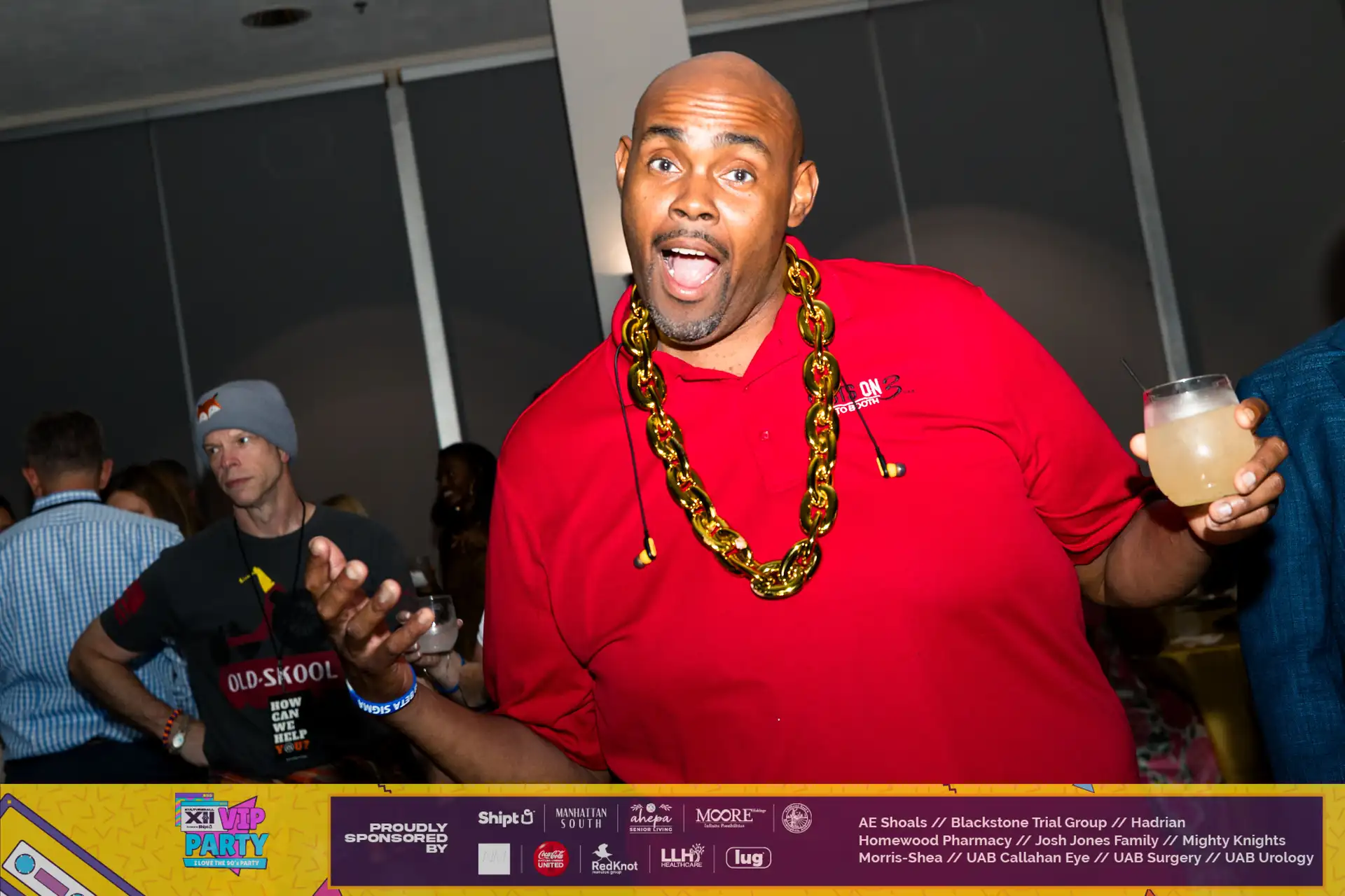 A man wearing a red shirt and a gold chain is standing in front of a group of people.