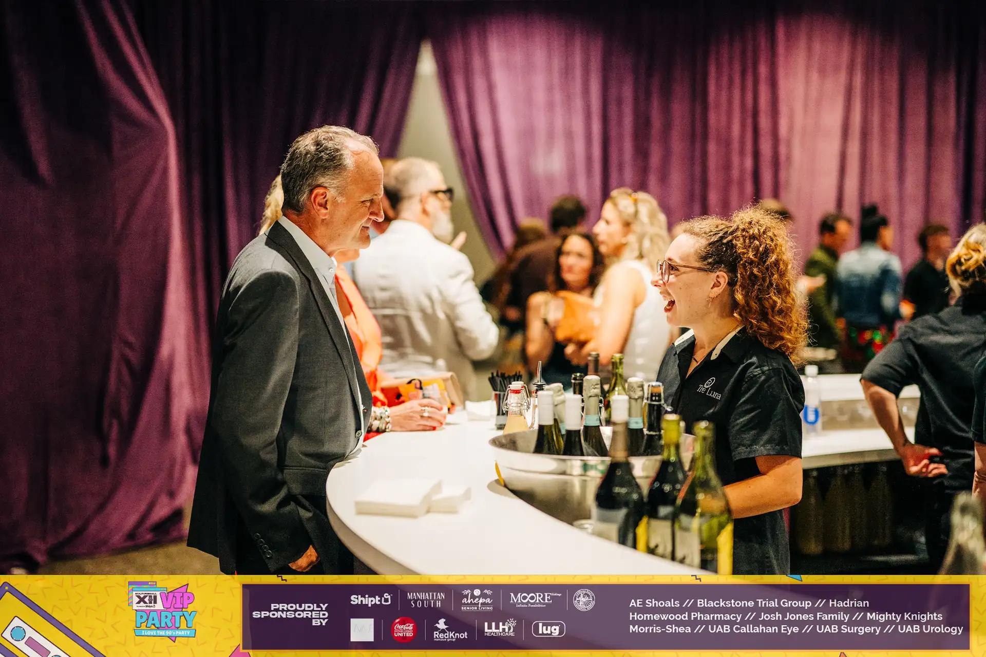 A woman behind a bar with a variety of wine bottles and glasses, serving drinks to guests at a high-end event.