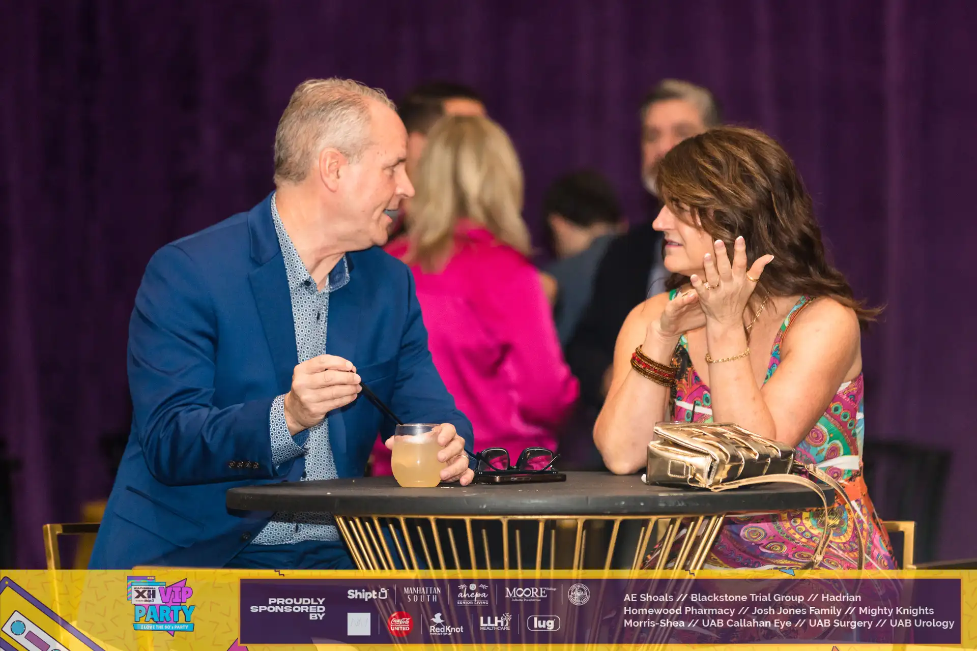 A man and woman sitting at a table with a drink in front of them, possibly at a high end event.