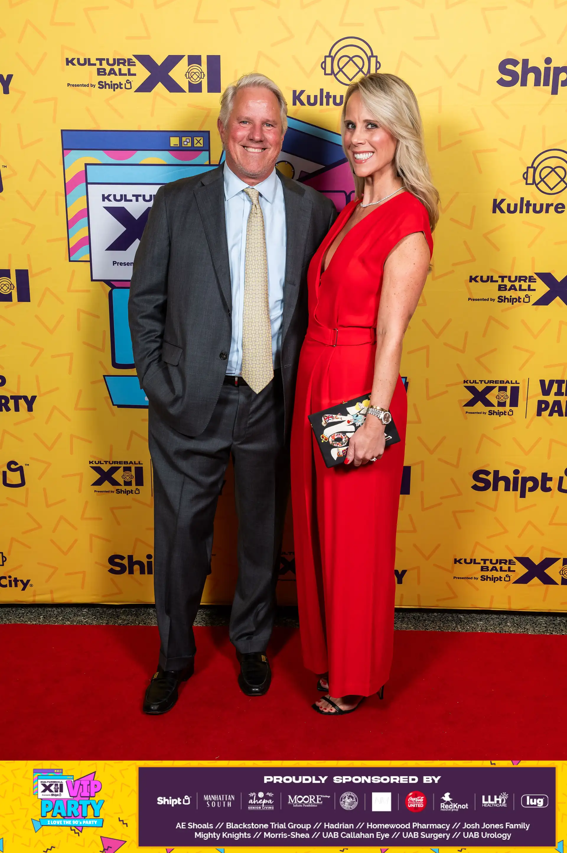 A man and woman standing on a red carpet, posing for a picture. The man is wearing a suit and tie, and the woman is wearing a red dress. They are in front of a yellow and blue backdrop, which has the words 'Kulture X' on it. The man is holding a bottle of Touch O Magic Lemonade.
