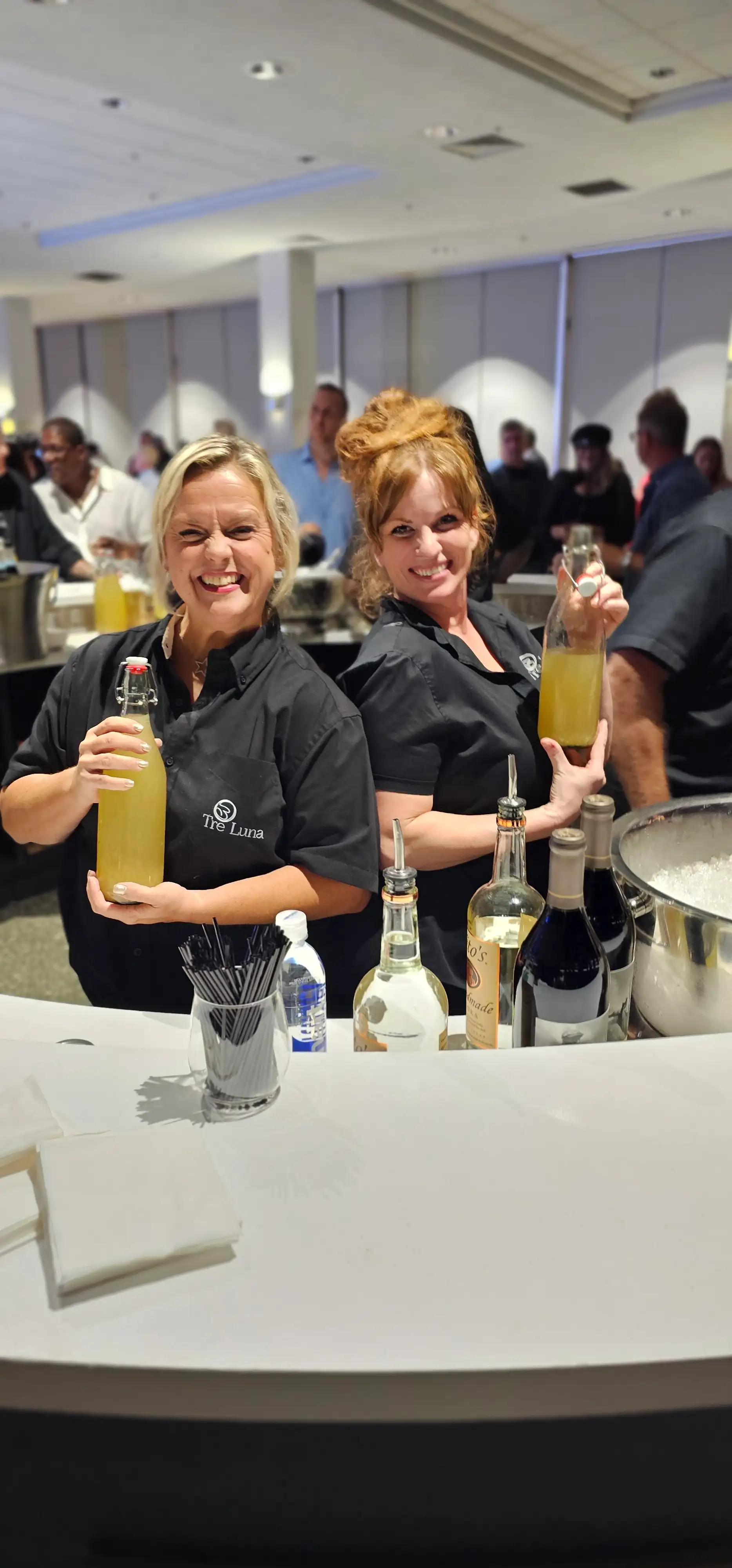 Two women in black shirts holding bottles of Touch O Magic Lemonade, a specialty drink for high-end events and catering services.