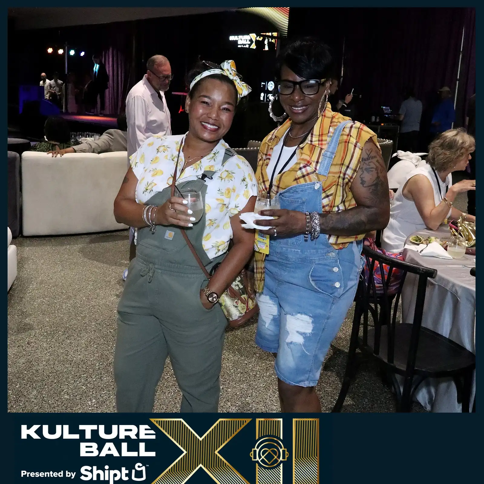 Two women in denim overalls holding drinks at a party.